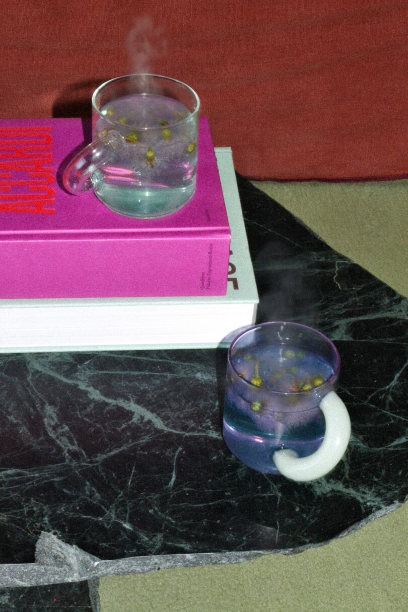 Two glass mugs with steaming herbal tea sit on a black marble table and a stack of books, one book with a bright pink cover