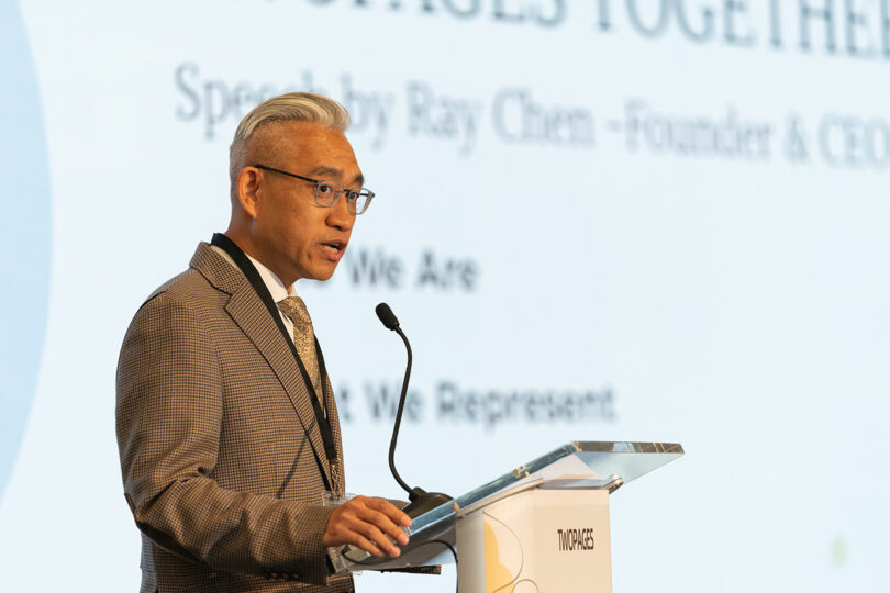A man in a suit and tie speaks at a podium with a microphone, presenting slides that highlight TWOPAGES behind him.