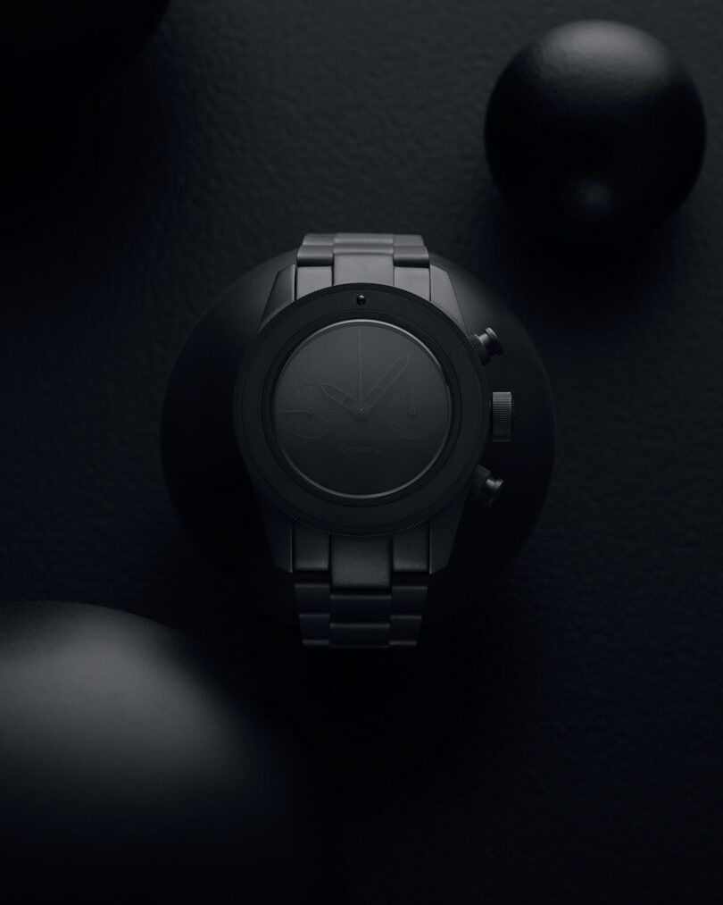 Matte black wristwatch with a metal band displayed on a dark textured background, surrounded by black spherical objects.
