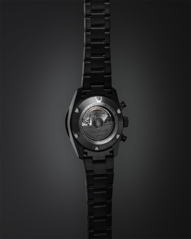 A black metal wristwatch with an exposed mechanical back, showing gears and movement, is displayed vertically against a dark gradient background.