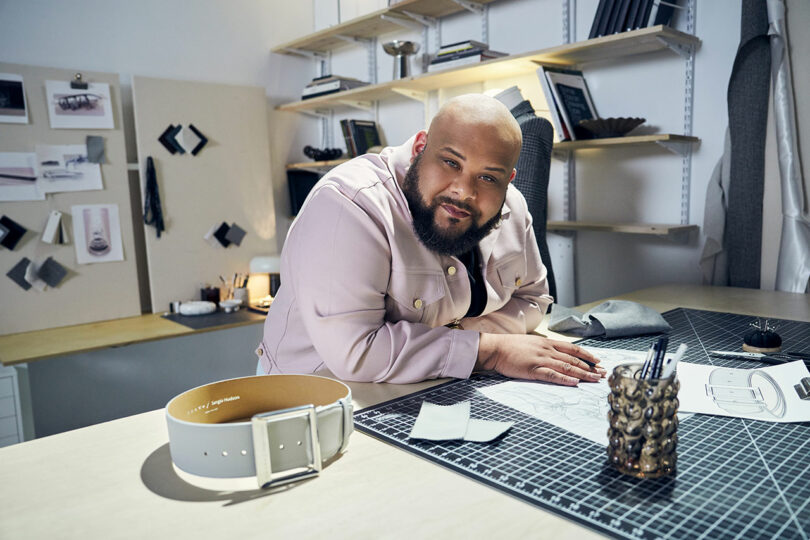 A person leans on a drafting table with sketches, fabric samples, and a belt, in a well-organized design studio.