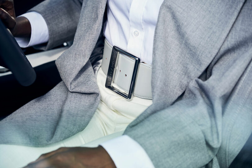 Person wearing a light gray suit jacket, white shirt, white pants, and a large silver-buckled belt while sitting in a car.