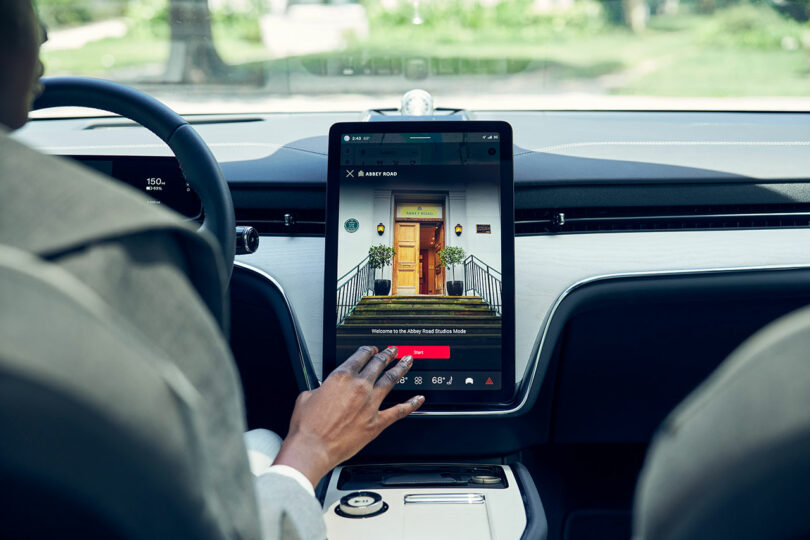 A person inside a car uses the large touchscreen display on the center console, which shows a photo of a house entrance with plants.