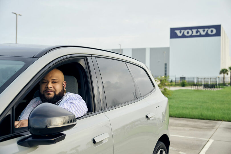A man sits in the driver's seat of a white SUV parked outside a Volvo building on a cloudy day.