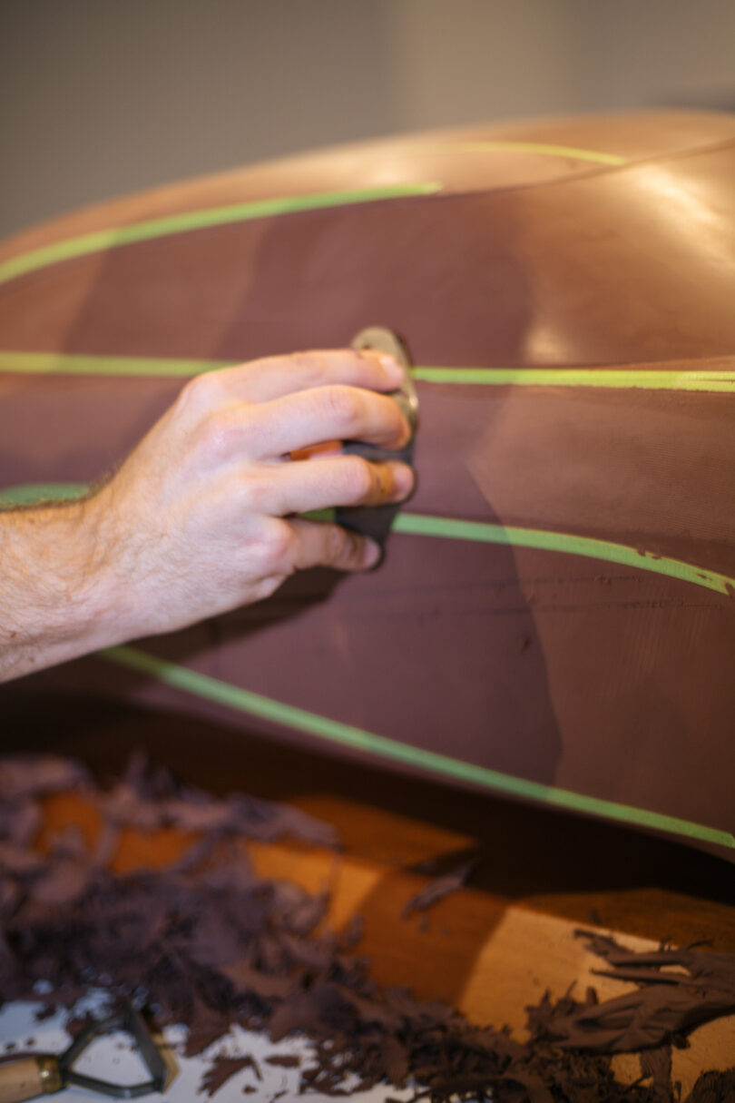 A person’s hand uses a tool to sculpt and smooth a brown surface with green lines, likely working on a model or prototype.