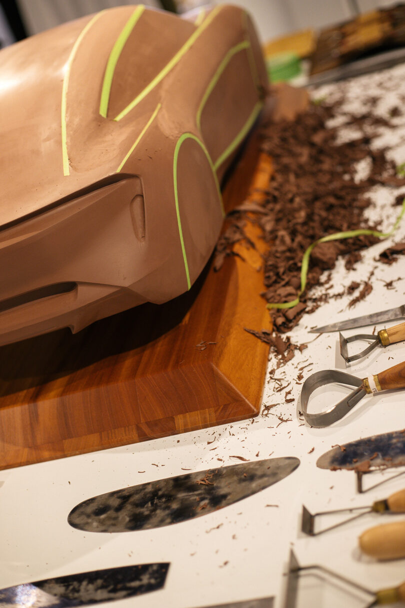 A clay model of a car in progress sits on a wooden base, surrounded by sculpting tools and shavings on a worktable.