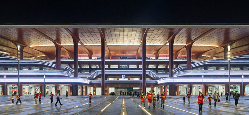 A modern building with illuminated horizontal lines at night; several people in red shirts are gathered and playing in the large open plaza in front.